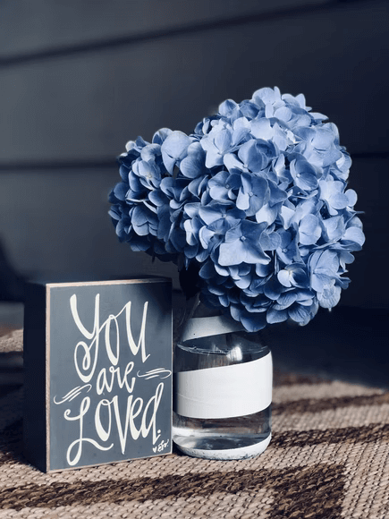 A small decorate block that says "You are loved" next to a blue hydrangea. 