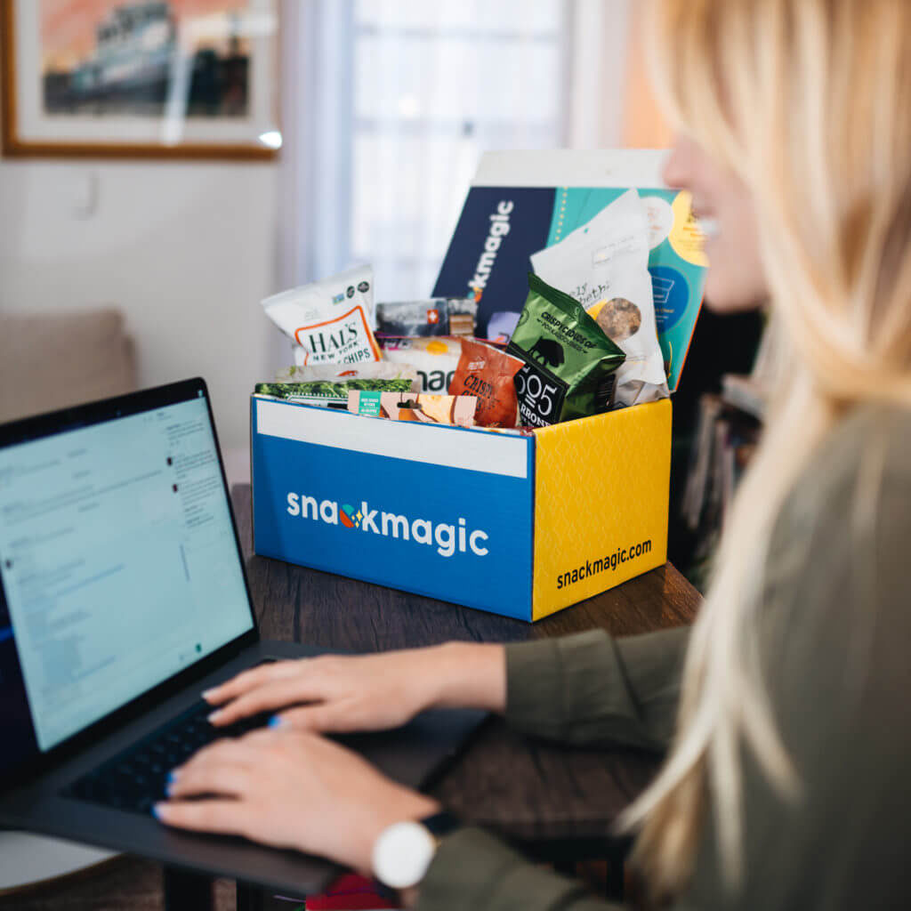 A virtual employee going through her emails on her laptop, sitting in her home office with a full SnackMagic box nearby. 