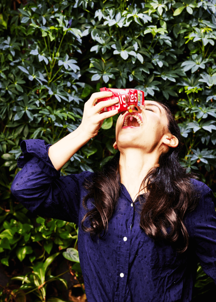 A woman standing next to a hedge, pouring a bagged snack enthusiastically into her mouth.