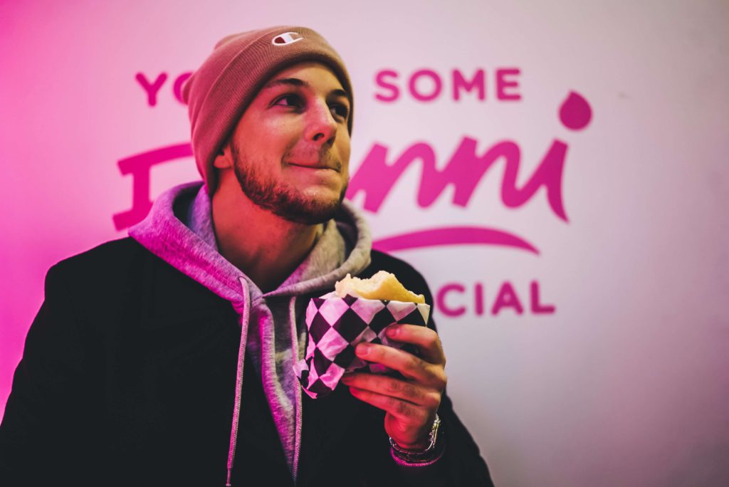 Young man enjoying a sandwich outside at night, lit up by neon lighting.