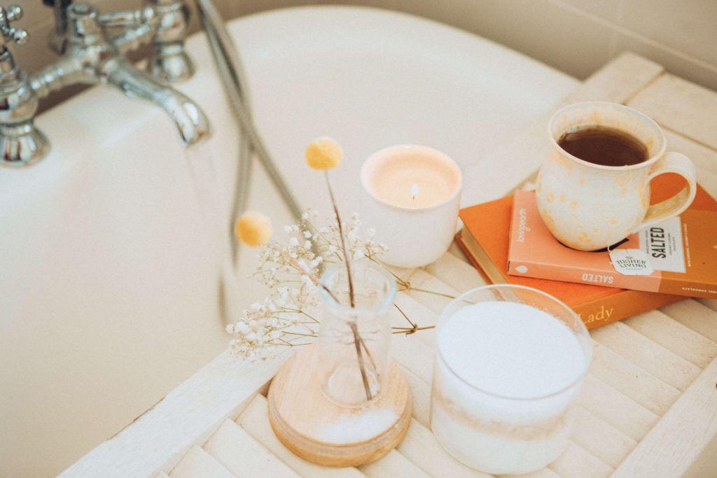 A self-care oriented gift set with tea, a candle, books, and flowers on a bathtub with running water. 