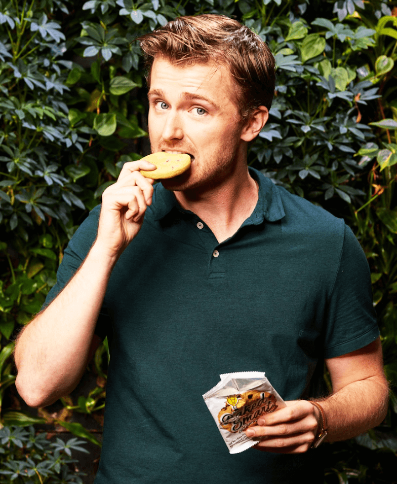 A young man in front of a hedge taking a bite of a cookie, one of his workplace snacks. 